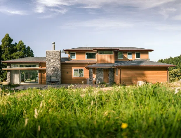 A contemporary multi-level timber-clad house set in a grassy landscape, with broad horizontal rooflines, extensive glazing, a prominent rough-stone chimney and entry surround, and warm wood wall cladding. The low, spread-out form is surrounded by mature trees and meadow vegetation.