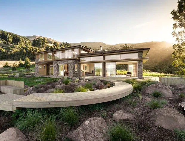Contemporary multi-level hillside residence with extensive floor-to-ceiling glazing, dark stone and timber-clad facade elements, and broad low-pitched rooflines. A curved timber walkway and deck-like platform wind through a landscaped rocky garden, with surrounding wooded hills and open mountainous terrain beyond.