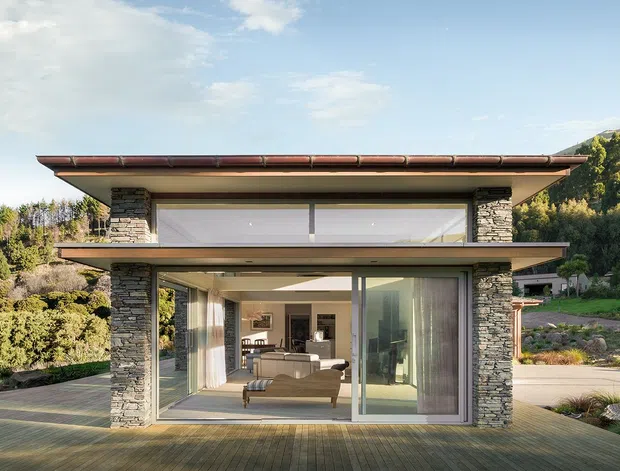A compact two-storey modern home set on a landscaped sloping site, with broad glazing, projecting flat roof planes, and stacked-stone side walls. The lower level opens onto a timber deck and reveals a bright living room, while the upper level features a long clerestory-style window. A wooded hillside and garden landscape surround the building.