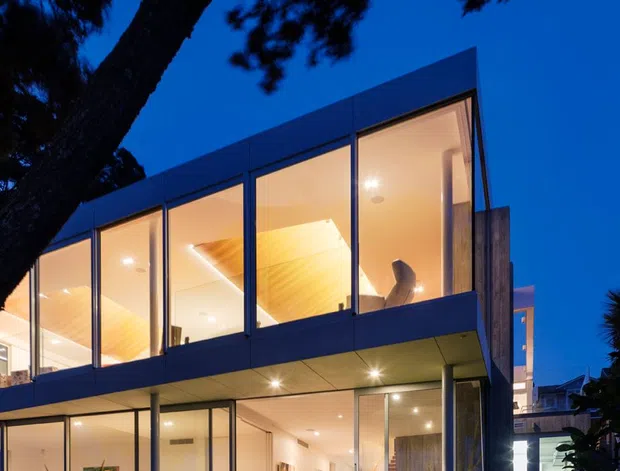 Twilight exterior view of a modern two-storey residence with extensive floor-to-ceiling glazing, dark framed windows, projecting upper-level volume, and illuminated open-plan interiors. A large mature tree and dense planting frame the house, while outdoor lounge furniture is visible beneath the cantilevered upper floor.
