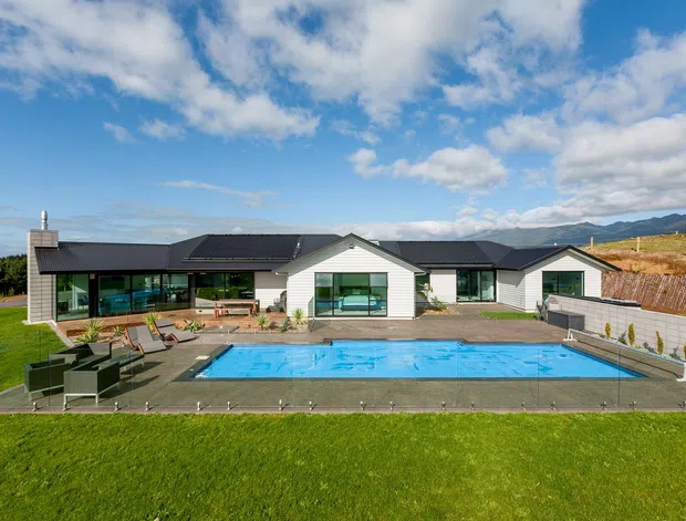 Wide exterior view of a low, single-storey modern residence with dark pitched roof sections, pale horizontal cladding, expansive glazing, and a central swimming pool surrounded by paved terraces and lawn. The property sits in an open elevated landscape with distant mountains visible beyond.