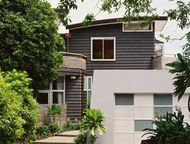 Exterior view of a compact multi-level contemporary house, largely screened by mature tropical and subtropical planting. The upper storey is clad in dark horizontal weatherboards with white-framed windows and a shallow low-pitched roof. A pale rendered garage volume projects in front, with a broad glazed garage door, while timber decking and curved balcony elements lead toward the entry.