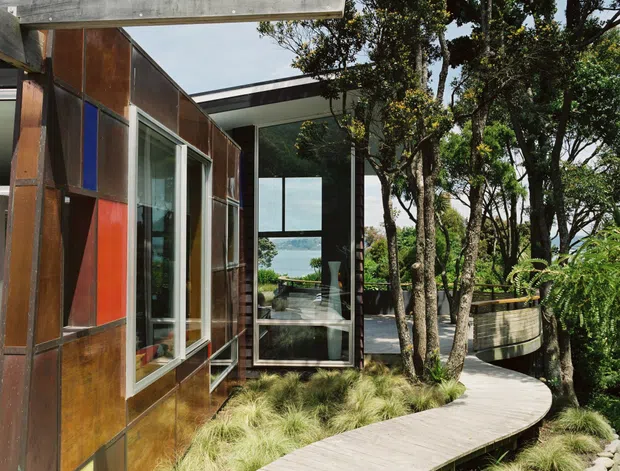Exterior view of a contemporary house set among mature trees, with weathered metal wall panels, large glazed openings, exposed concrete beams, and a curved timber walkway through ornamental grasses and stones. Water is visible through the glazing in the background.