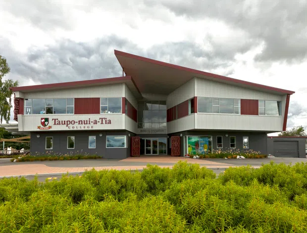 Exterior view of a contemporary two-storey school building with a prominent asymmetrical flat roof and deep projecting canopy. The facade combines light grey metal cladding, dark grey lower walls, red accent panels, extensive horizontal glazing, and a central recessed glazed entrance. Landscaping and a paved forecourt sit in front of the building, with the school name visible on the facade.