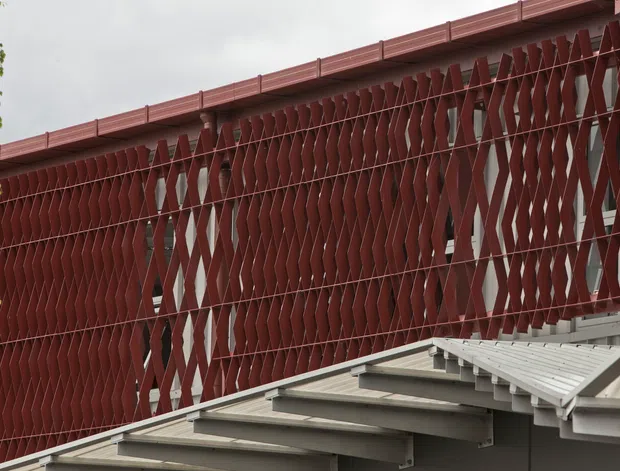 Close-up exterior architectural detail showing a red-brown perforated and folded metal screen facade with a repeating diamond-like geometric pattern. A low sloping roof with exposed grey structural framing occupies the foreground, while sparse greenery and an overcast sky appear at the edges. The image emphasizes facade screening, roof construction, and industrially detailed exterior materials.