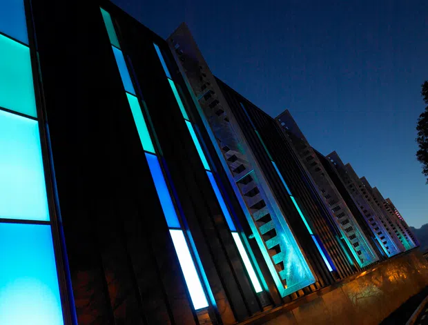 Dramatic low-angle nighttime view of a large contemporary multi-storey building facade. Repetitive vertical bays combine dark cladding, illuminated turquoise, blue and white glazing or light panels, and projecting concrete or metal structural elements. Warm exterior lighting grazes the lower wall, creating a high-contrast, futuristic architectural composition.