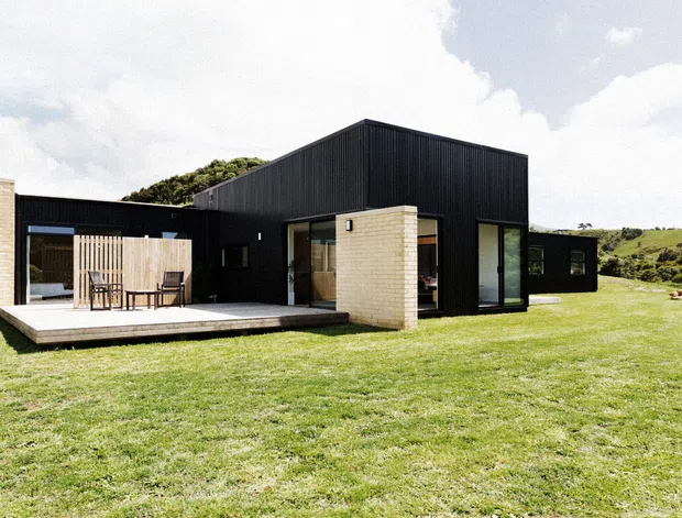 A low, modern black-clad house set in an expansive grassy landscape. The building combines crisp rectilinear volumes with pale brick feature walls, large glazed openings, a timber outdoor deck, and a partially enclosed courtyard-like entertaining area with outdoor dining furniture. Rolling green hills form the distant background.