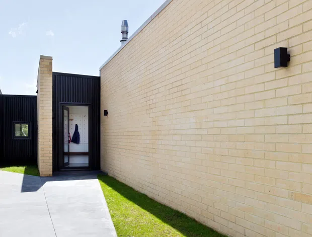 Exterior view of a contemporary residential entry approach, featuring a long pale cream brick wall, dark vertical metal cladding, a recessed glazed entry door, concrete paving, and a narrow strip of lawn under bright daylight.