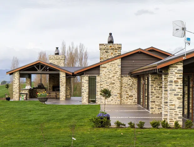 Single-storey residential house set in a rural landscape, featuring extensive irregular natural-stone masonry walls and chimneys, dark horizontal timber cladding, and multiple pitched gabled roof forms with wide eaves. A covered outdoor entertaining area with a fireplace is visible at left, while paved paths and manicured lawn surround the building. The design has a rustic contemporary lodge or farmhouse character.