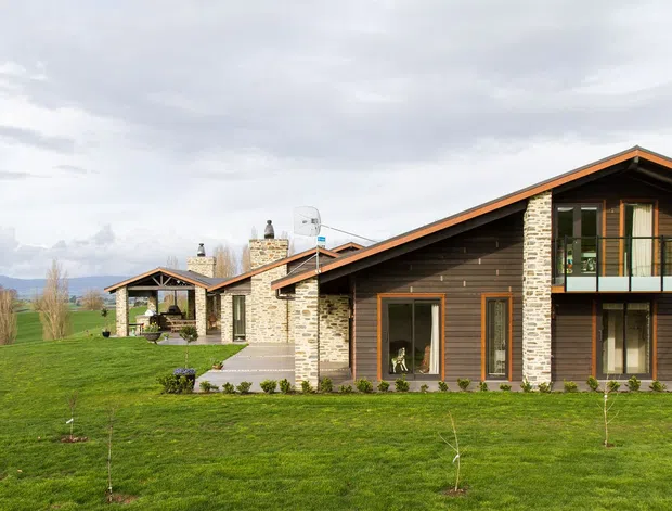 A contemporary single-storey-plus-mezzanine rural house with a long pitched gable roof, dark horizontal timber cladding, and prominent pale stone masonry walls and chimney elements. Large glazed openings and a covered terrace face a broad green lawn, with distant mountains and open countryside visible beyond.