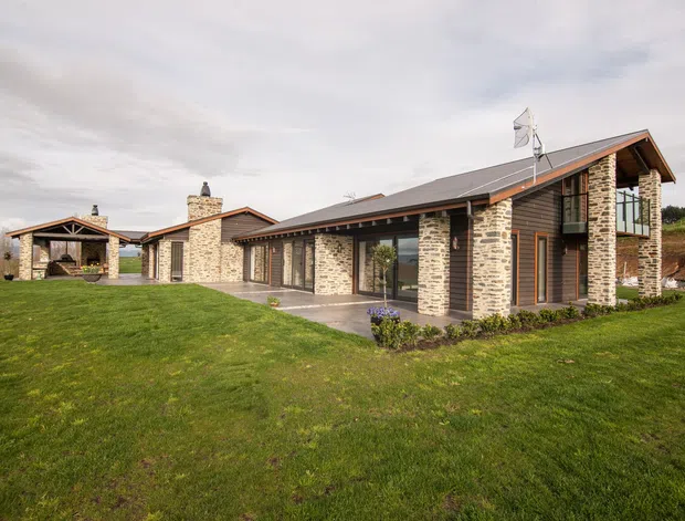 A low, elongated residential building set in a broad grassy landscape. The exterior combines pale rough stone masonry, dark horizontal timber cladding, and warm timber roof and window trims beneath a long pitched roof with deep eaves. A covered outdoor structure and stone chimney are visible at the left, while the main facade has large glazed openings and sheltered outdoor circulation. The setting appears rural and open, with gently sloping terrain and an overcast sky.