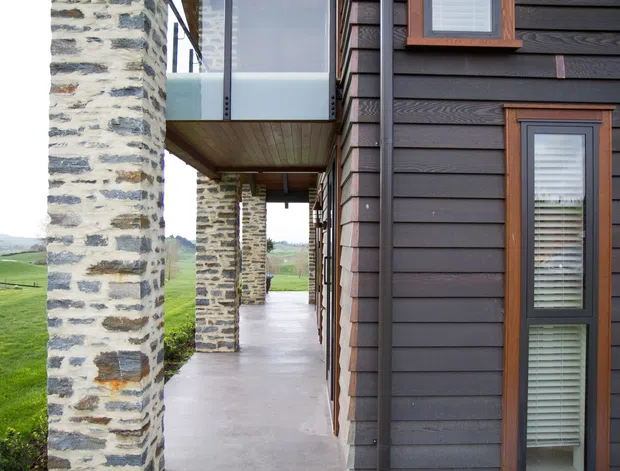 Close exterior architectural view of a contemporary rural home featuring dark horizontal timber cladding, warm timber-framed windows, exposed natural stone masonry columns, and a covered concrete walkway. Glass balustrading and an upper-level connection are visible, with rolling green farmland beyond.