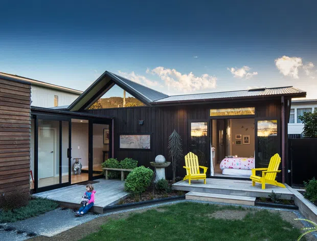 Exterior view of a compact modern dark timber-clad house or extension arranged around a small landscaped courtyard. The building has a prominent pitched gable roof, large glazed doors and windows, a covered entry, raised timber deck, and bright yellow outdoor Adirondack chairs. Low planting, lawn, gravel paths, and neighbouring structures are visible.