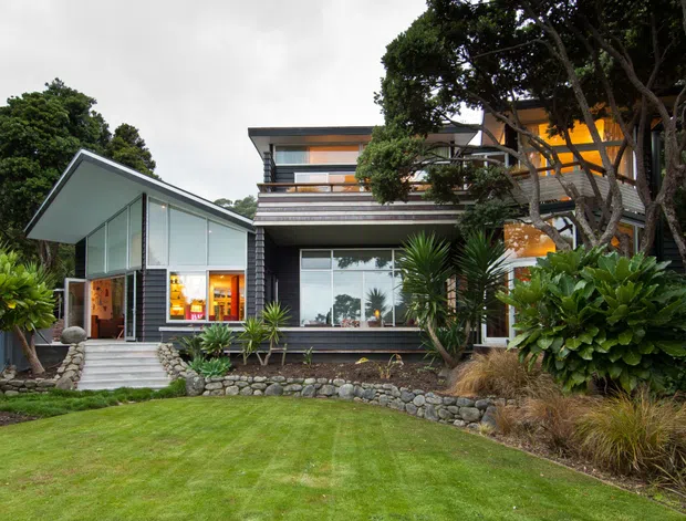 Exterior view of a multi-level contemporary home set within a landscaped garden. The house features strong horizontal lines, extensive floor-to-ceiling glazing, dark horizontal cladding, projecting roof forms, layered balconies, and warm interior lighting visible through the windows. A manicured lawn, stone retaining edge, palms, and established trees surround the building.