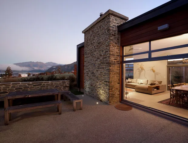 A dusk exterior view of a contemporary home with a low-profile roofline, large floor-to-ceiling glazed doors, dark timber cladding, and a prominent rough stone chimney or masonry wall. A paved terrace with built-in stone planter walls and timber picnic-style seating overlooks a mountainous landscape. The warmly lit interior visible through the glazing contains a living room and dining area.