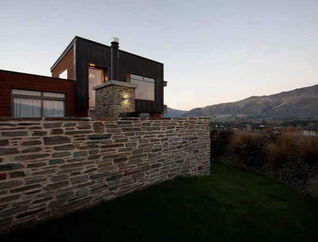 Exterior view of a contemporary multi-level home at dusk, featuring dark metal cladding, warm timber-clad walls, large glazing, a prominent stone chimney, and a long dry-stone boundary wall. The property sits in a landscaped setting with mountain terrain and neighbouring rooftops visible beyond.