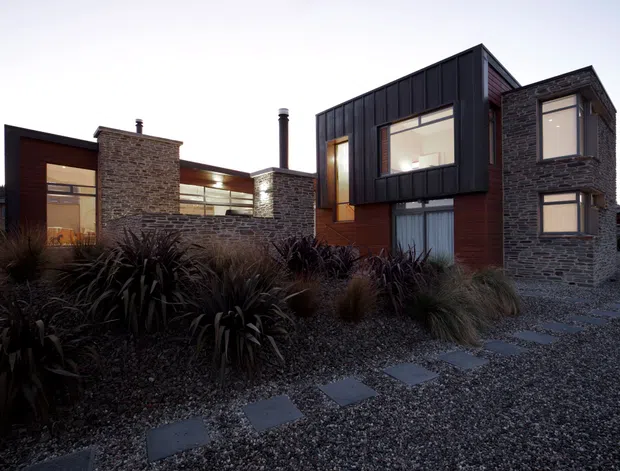 Exterior view of a contemporary, multi-level house composed of dark stone masonry, vertical dark metal cladding, and warm timber wall panels. Large windows and glazed openings connect the interior to a low-maintenance gravel landscape with native-style grasses and stepping stones. The building has varied cubic volumes, projecting upper-level forms, and tall chimney elements.