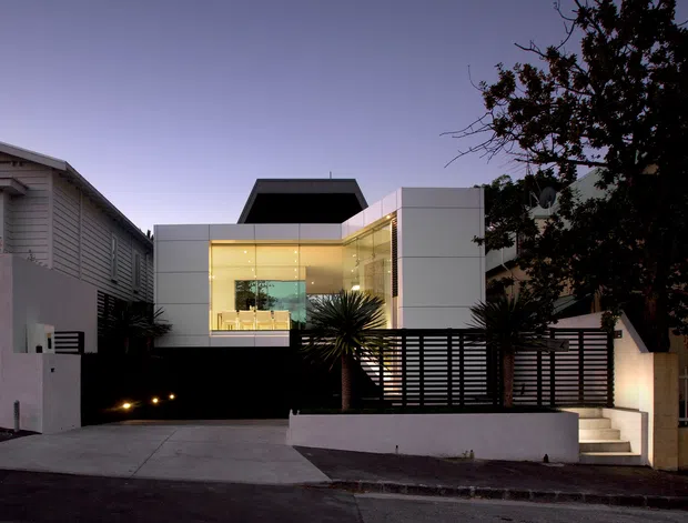 Twilight street view of a contemporary multi-level residence with a crisp white geometric facade, extensive illuminated corner glazing, dark roof forms, horizontal slatted screening, a concrete driveway, and stepped entry landscaping. Adjacent suburban buildings and mature trees frame the property.