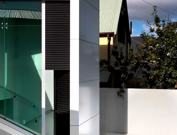 A contemporary residential exterior showing a sharply geometric white-clad entry volume with tall green-tinted glazing, dark horizontal louvres, and a glass stair balustrade. A curved path of round concrete stepping stones leads through a minimalist gravel courtyard. An older pitched-roof house is visible behind the modern addition.