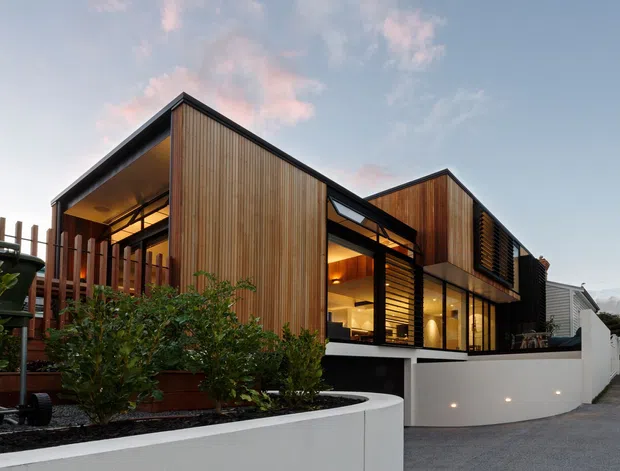 Exterior view of a contemporary two-storey residential building with sharply angled pitched roof forms, warm vertical timber cladding, dark window and door frames, and extensive glazed openings. Curved white rendered retaining walls and landscaped planting define the foreground, while warm interior lighting creates an inviting evening appearance.