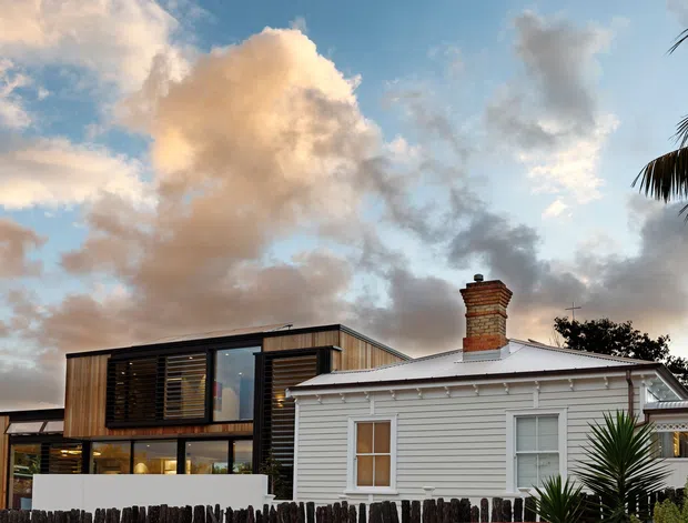 Exterior view of a renovated home combining a traditional white weatherboard cottage with a contemporary timber-clad addition. The cottage has a low-pitched metal roof, brick chimney, horizontal cladding, and white-framed windows, while the modern extension features dark timber screening, large glazing, and clean rectangular forms. Palm and other planting frame the scene, with a partly cloudy sunset sky in the background.