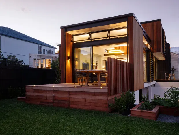 Twilight exterior view of a contemporary timber-clad residential extension with a glazed living and dining space opening onto a raised timber deck. The design features strong dark framing, warm interior lighting, vertical timber screening, built-in deck planters, and a flat roof. An adjacent older house and church-like spire are visible in the background.
