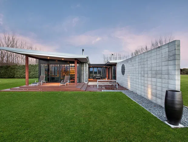 Single-storey contemporary pavilion-style home set in a broad manicured lawn, with low-slung sloping metal roofs, extensive glazing, timber decking and posts, and a prominent pale grey concrete block privacy wall. The outdoor terrace and glazed living spaces face the lawn, while gravel landscaping borders the wall.