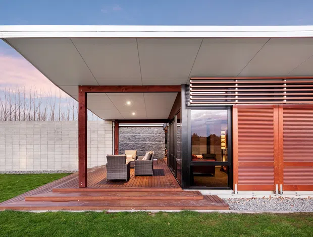 A low-profile modern pavilion with a broad flat roof, deep covered outdoor living area, timber decking, and floor-to-ceiling glazing. The facade combines dark framing, warm timber-clad sliding screens, horizontal louvres, and pale masonry boundary walls. Outdoor lounge furniture sits beneath the covered terrace, with a neatly landscaped lawn in the foreground.