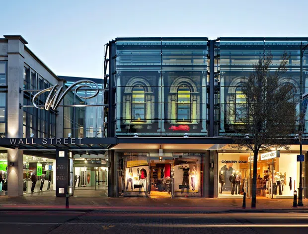 Street-level view of a contemporary urban retail and commercial building, with illuminated shopfronts at ground level and a prominent glazed upper facade incorporating large arched windows. The frontage includes the visible 'Wall Street' sign, sculptural metal canopy elements, dark framing, and adjacent multi-storey commercial facades.