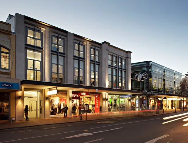 Twilight streetscape showing a contemporary multi-storey commercial retail building with tall framed glazing, pale stone or concrete facade panels, projecting canopies, illuminated shopfronts, and a glazed corner structure. The image is photographed from across a multi-lane urban street, with vehicle light trails in the foreground and adjacent retail buildings along the block.