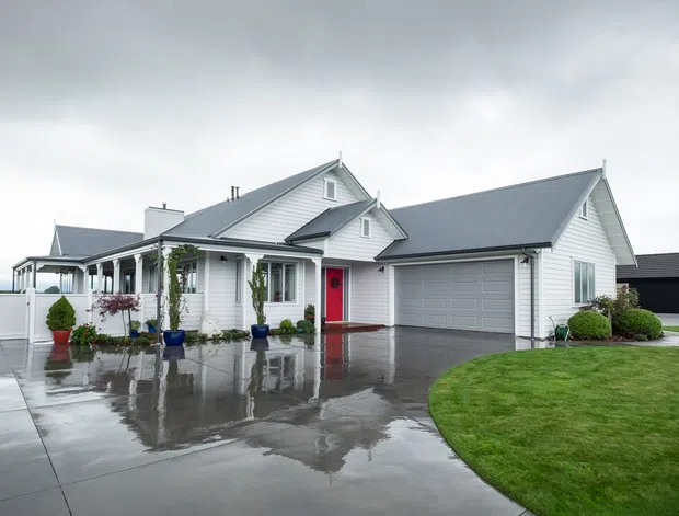 Exterior photograph of a large white weatherboard-style suburban home in the rain, featuring multiple pitched gabled roofs, a covered front veranda with white columns, a bright red front door, grey garage door, and broad wet concrete driveway. Neatly landscaped lawn and potted plants surround the entrance.
