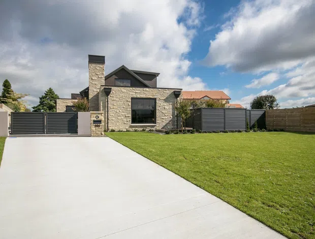 Street-facing view of a contemporary single-storey house with a dark pitched roof, pale stacked-stone facade and chimney, large dark-framed window, gated driveway, lawn, and horizontal privacy fencing. A neighbouring terracotta-roofed home is visible behind the property.