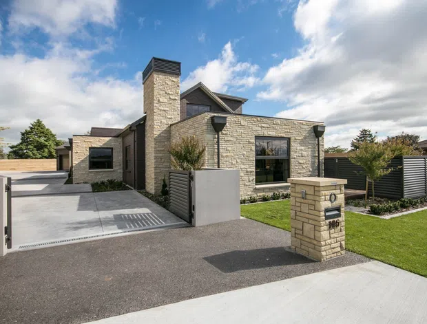 Street-facing exterior view of a contemporary single-storey home with pale stacked-stone cladding, a prominent stone chimney, dark-framed windows, low-pitched roof forms, landscaped lawn, driveway, and a stone letterbox at the front boundary.