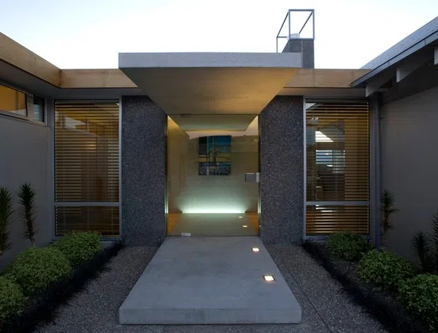 A symmetrical modern residential entrance courtyard viewed from outside. A broad concrete path leads beneath a projecting flat-roofed canopy into a recessed glazed entry, flanked by dark textured masonry or concrete walls and tall windows with horizontal timber blinds. Gravel landscaping, clipped shrubs and narrow upright plants line both sides, while small recessed path lights illuminate the approach. The composition is geometric, restrained and strongly horizontal.