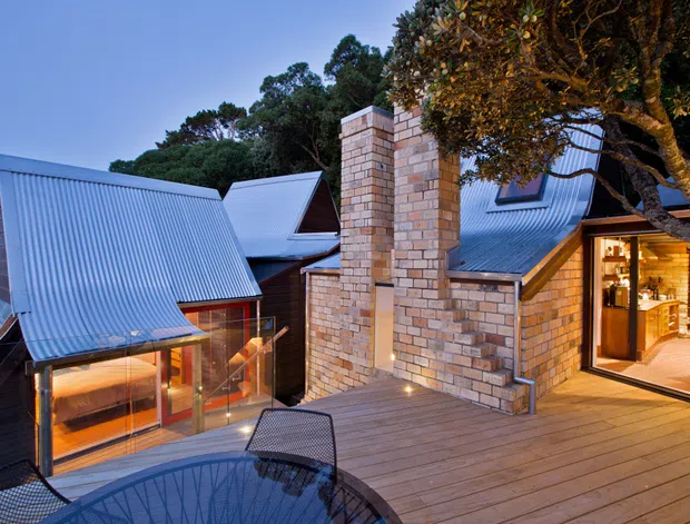 A dusk exterior view of a multi-level contemporary residential building set among mature trees. The design features steeply pitched corrugated metal roofs, warm-toned brick chimney-like masonry walls, extensive glazing, timber detailing, and a broad timber deck with outdoor furniture. Warm interior lighting is visible through the glazed doors and windows.