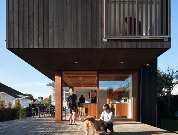 A contemporary two-storey residential house photographed from the rear, with a dramatic cantilevered upper volume clad in dark vertical timber boards. The ground floor opens onto a broad timber deck through large glazed doors, revealing a warm timber-lined interior and kitchen. Vertical timber screening forms a balcony or upper-level opening, while the deck and garden create an outdoor living area. People and a dog are present but are secondary to the architecture.