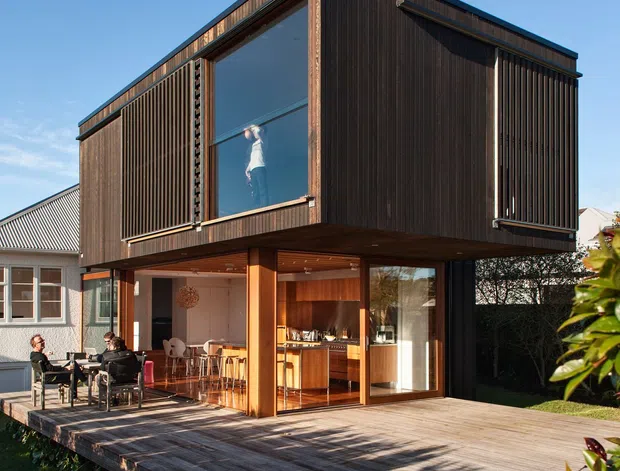 A contemporary two-storey residential extension viewed from the garden, featuring a projecting upper-level box clad in dark vertical timber slats, large glazed openings, and dark metal trim. The ground floor opens onto a broad timber deck with an outdoor dining area and a warm timber-lined kitchen visible inside. The extension is raised above the deck on timber-clad supports and connected to an older pitched-roof house.