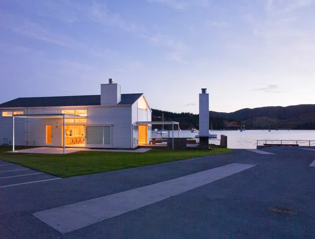 Dusk exterior view of a low-rise modern white-clad building beside a waterfront. The building has a simple gabled roof, projecting covered outdoor areas, large glazed openings with warm interior lighting, and prominent white chimney forms. A paved forecourt and lawn lead toward the water, with wooded hills visible across the inlet.