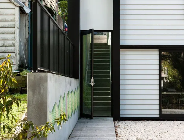 Exterior view of a contemporary residential entry passage between two weatherboard-clad structures. A tall black-framed glazed door sits within a projecting black portal, opening onto a narrow timber walkway. White horizontal cladding, dark metal fencing, a rendered boundary wall with a geometric green mural, gravel, and concrete paving define the composition.