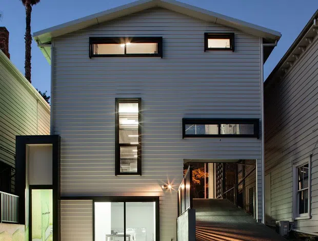 Twilight exterior view of a narrow multi-storey contemporary house in an urban infill setting. The facade has horizontal light-coloured weatherboard cladding, a simple gabled roof, dark-framed rectangular windows, and a glazed ground-floor opening. A steep driveway or passage runs alongside the house, with neighbouring buildings tightly framing the site and a palm tree visible at left.