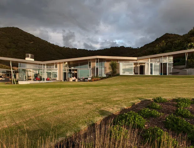 A low-profile single-storey modern house set within a broad landscaped lawn and surrounded by densely vegetated hills. The building has extensive floor-to-ceiling glazing, slender light-coloured structural framing, a long shallow roof plane with broad overhangs, and transparent indoor-outdoor living areas. The scene is photographed under dramatic overcast skies with warm sunlight catching the facade.