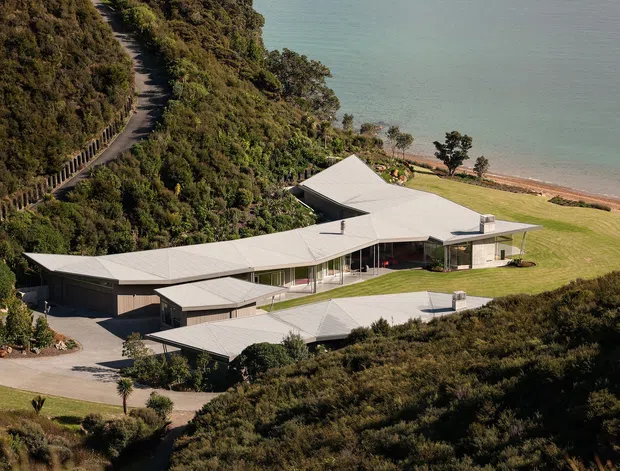Aerial view of a sprawling contemporary coastal residence set into a vegetated hillside above a beach and calm body of water. The single-storey home has multiple interconnected wings, broad low-pitched light metal roofs, extensive glazing, dark exterior walls, and landscaped lawn areas. A narrow hillside access road and shoreline are visible.