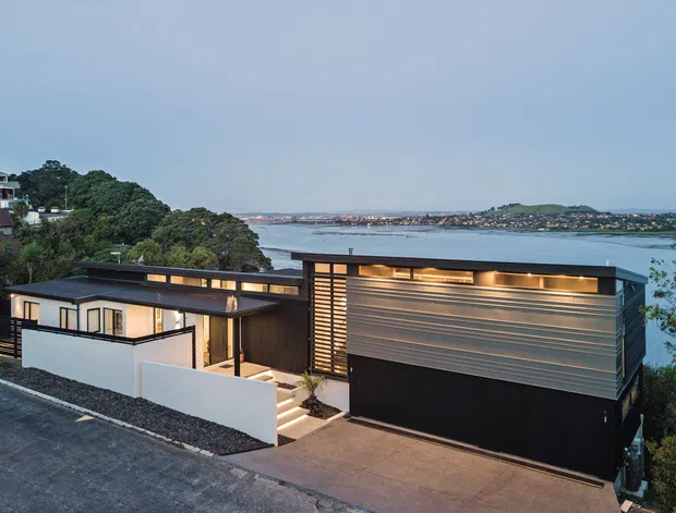 A contemporary multi-level waterfront residence viewed from outside at dusk. The house features a dark flat-roofed facade, horizontal metal cladding, extensive glazing, warm linear lighting, white rendered privacy walls, and a stepped driveway and entry. Water, distant shoreline development, and landscaped hills are visible beyond the property.