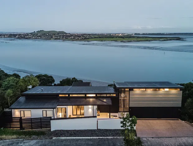 Aerial view of a contemporary waterfront residence with dark metal pitched and low-slope roof forms, horizontal timber-like screening, pale rendered walls, and expansive glazed openings. The home sits above a broad tidal inlet with a distant developed shoreline and landscaped vegetation around the property.