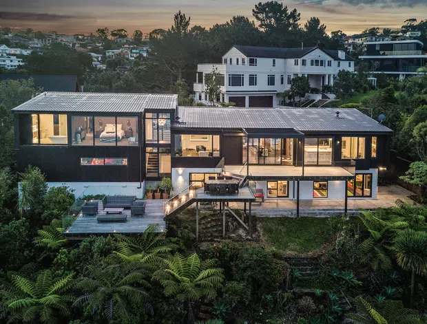 A dusk aerial view of a contemporary multi-level house set into a lush, densely planted hillside. The home features dark cladding, extensive glazing, low-pitched corrugated metal roofs, multiple decks and terraces, and warm interior lighting. Surrounding suburban homes and mature vegetation are visible in the background.