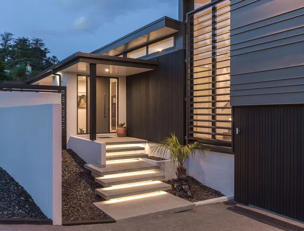 Contemporary residential facade at dusk, featuring strong horizontal lines, dark metal or timber cladding, a projecting entry canopy, illuminated floating concrete steps, vertical timber screening, and a large dark garage door. Low-maintenance gravel landscaping and a partially enclosed entry courtyard frame the approach.