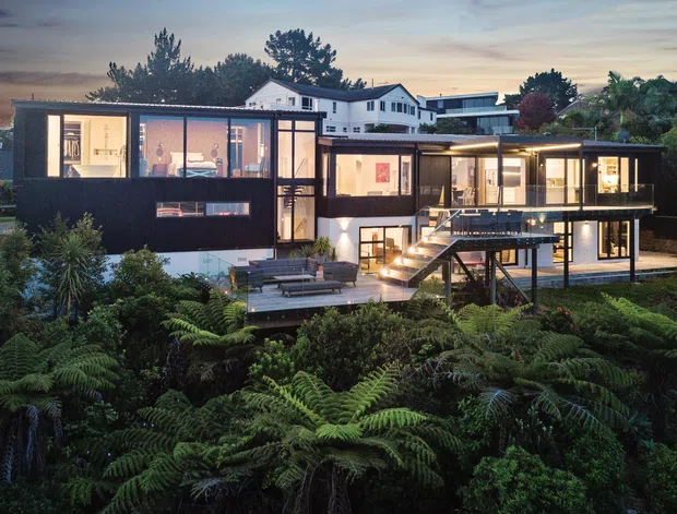 Twilight exterior view of a large contemporary multi-level home set within dense lush vegetation. The house features dark cladding, extensive floor-to-ceiling glazing, projecting forms, multiple terraces and decks, and a prominent exterior stair connecting the landscaped garden to the upper living areas. Warm interior lighting contrasts with the dark facade and surrounding greenery.