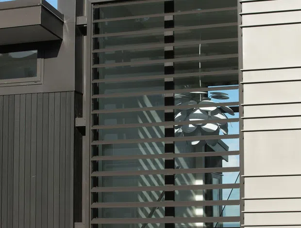 Close-up exterior view of a modern multi-level residential facade with a large glazed stairwell or entry volume screened by horizontal metal louvres. Dark vertical cladding, pale horizontal wall cladding, charcoal trim, and projecting flat roof forms create a strong contemporary composition. Interior stairs and a sculptural pendant light are visible through the glazing.