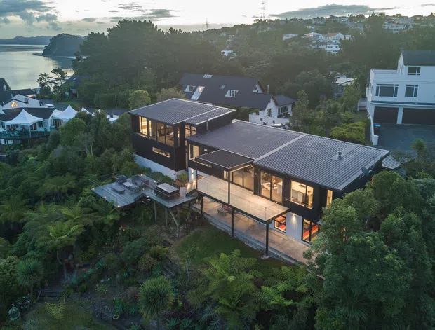 Aerial view of a contemporary multi-level home set into a lush, steeply vegetated coastal hillside. The dark-clad building has two connected pavilion-like wings with low-pitched metal roofs, extensive floor-to-ceiling glazing, warm interior lighting, and elevated decks or platforms. A bay or inlet is visible to the left, with neighbouring homes distributed across the wooded hillside.
