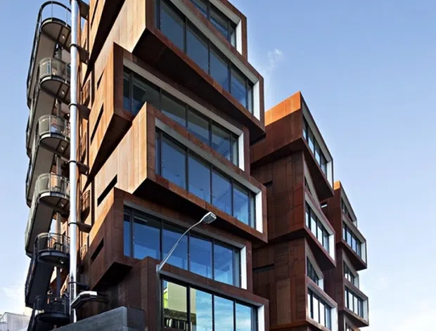 Street-level exterior view of a contemporary multi-storey urban apartment or mixed-use building. The facade combines extensive floor-to-ceiling glazing with projecting, irregularly stacked bays clad in dark rust-brown metal or weathered steel, contrasted by exposed grey concrete at the lower level. Curved external balconies and services are visible along the left side, with adjacent urban buildings and a street setting.
